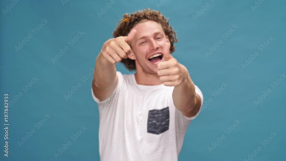 Cheerful curly man in t-shirt showing thumbs up at the camera over blue background Cheerful curly man in t-shirt showing thumbs up at the camera over blue background