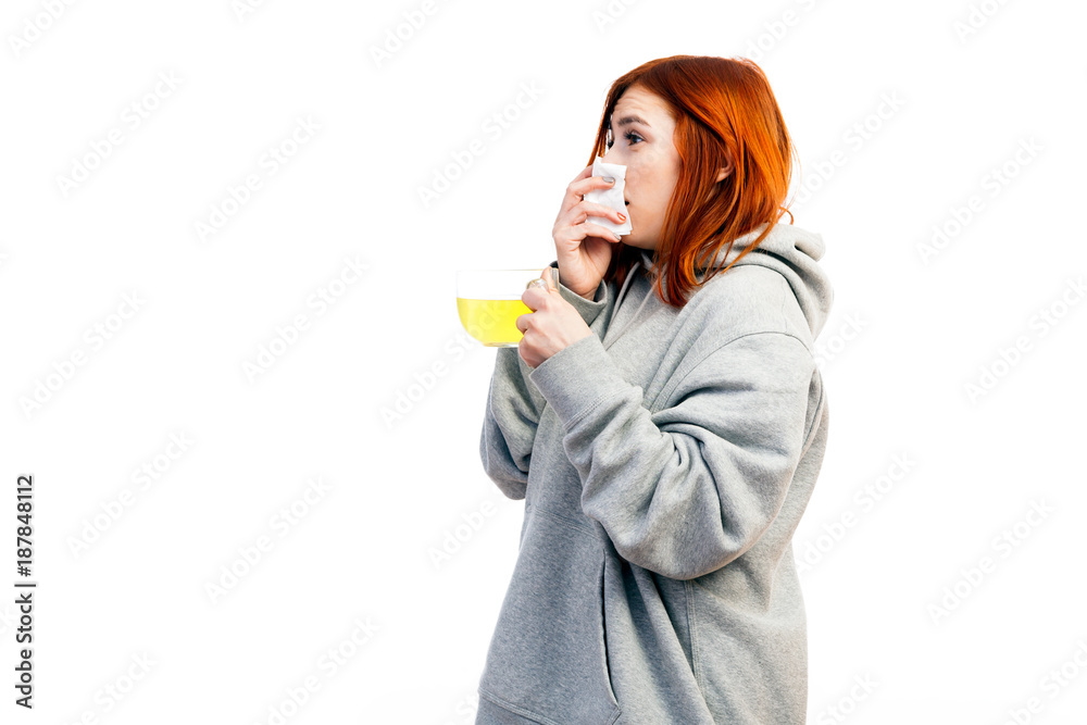 A young red-haired woman in a gray sweatshirt is sick, sneezes and drinks from a large transparent cup of an antiviral agent for treatment on a white isolated background A young red-haired woman in a gray sweatshirt is sick, sneezes and drinks from a large transparent cup of an antiviral agent for treatment on a white isolated background