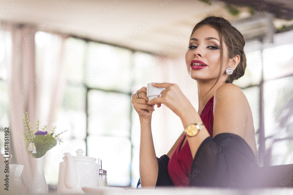 Beautiful brunette caucasian business woman sitting in cafe on her coffee break in red dress a bit classic dress and black cardigan, smiling. Copy space. Rich luxury place. Evening, sunset light