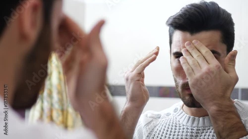 Handsome young man applying moisturizing cream on face in front of home bathroom mirror