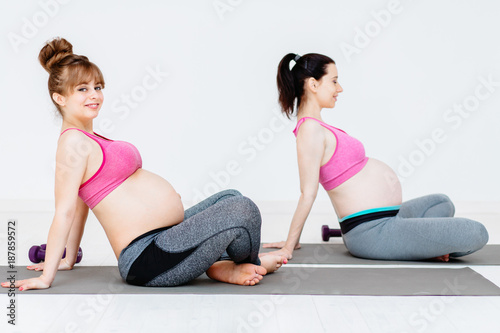 Sport, maternity and liesure concept - two pregnant women relaxing and stretching their back after workout at home or yoga class.