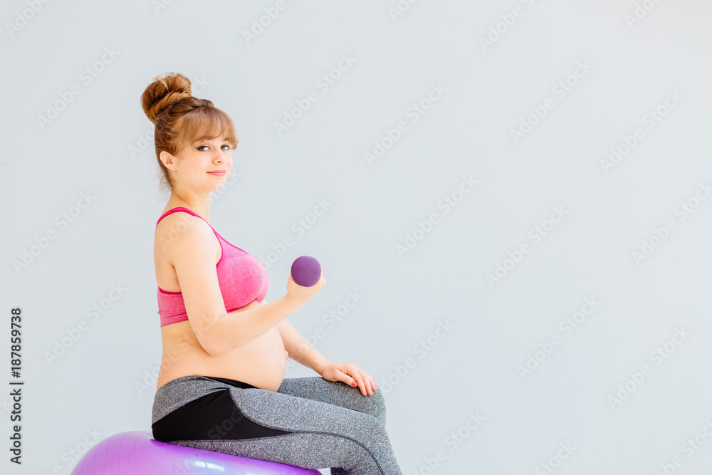 Pregnant woman with bun hairstyles in sportswear is doing exercises with dumbbell while sitting on gymnastic ball. Pregnant woman with bun hairstyles in sportswear is doing exercises with dumbbell while sitting on gymnastic ball.