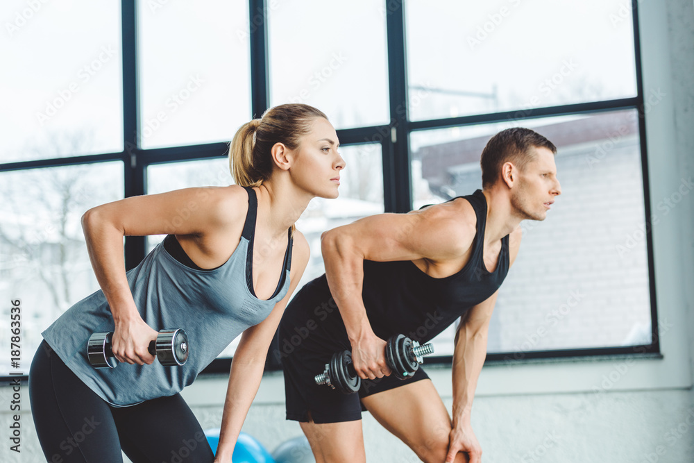 side view of couple in sportswear training with dumbbells in gym side view of couple in sportswear training with dumbbells in gym