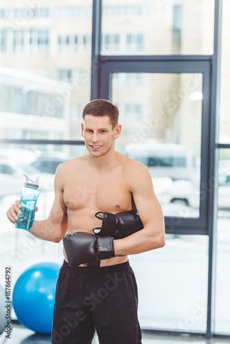 young muscular sportsman in boxing gloves drinking water from bottle and looking at camera in gym