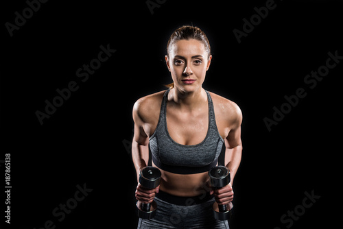 athletic young woman in sportswear exercising with dumbbells and looking at camera isolated on black