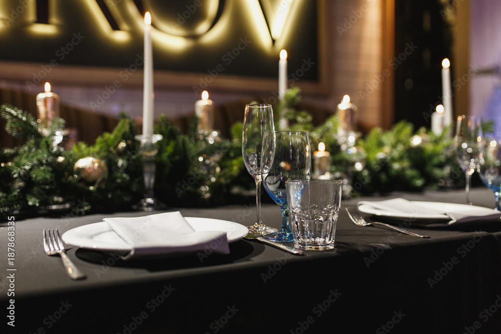 Close up glasses, forks, knives, napkins, candles and decorative flower on dark black tablecloth on table served for dinner in cozy restaurant. Empty served restaurant table with plates and wine glass Close up glasses, forks, knives, napkins, candles and decorative flower on dark black tablecloth on table served for dinner in cozy restaurant. Empty served restaurant table with plates and wine glass