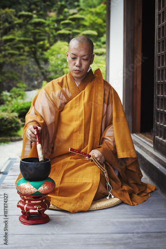 Buddhist monk with shaved head wearing golden robe sitting on floor outdoors, using singing bowl. photo