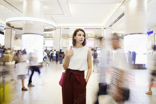 Young woman with brown hair wearing white sleeveless top and red trousers standing in shopping mall, blurred motion of passers by.