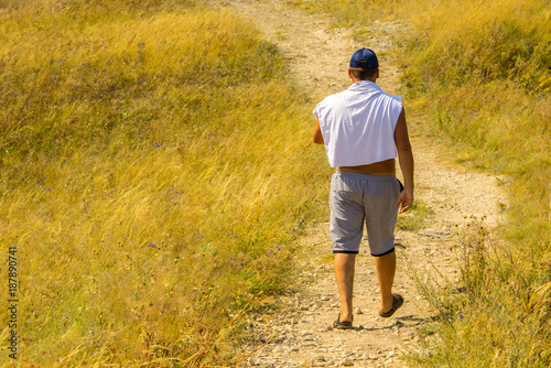 A man in shorts, a white T-shirt and a blue cap walking along a sandy and stony path among bushes, grass and flowers. © Dmitrii