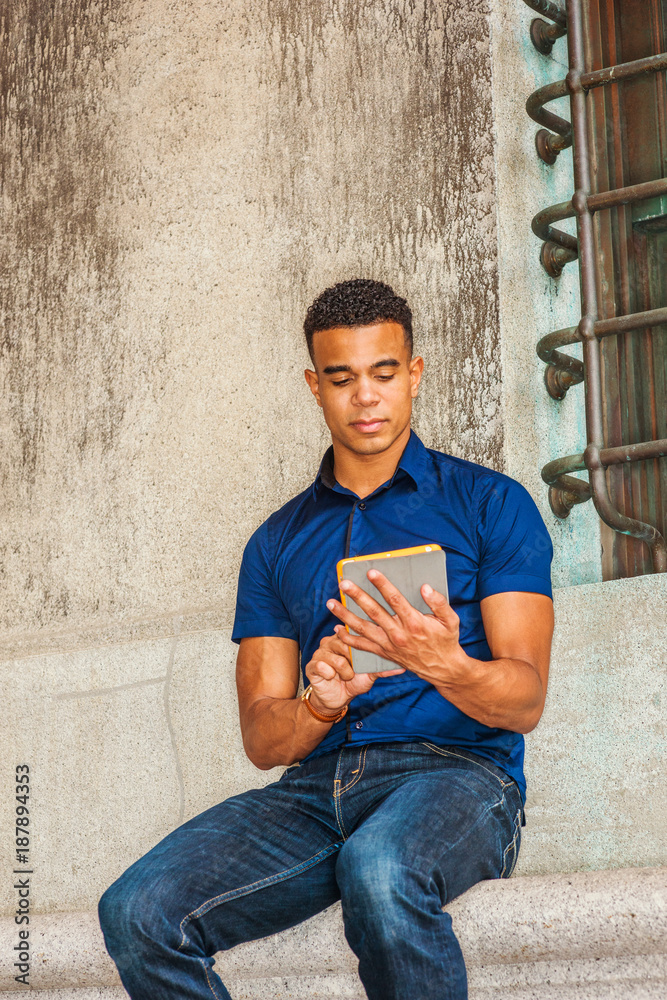 I love reading. Modern Reading. Wearing blue short sleeve shirt, jeans, African American college student sitting against vintage wall on street in New York, reading small tablet computer. . I love reading. Modern Reading. Wearing blue short sleeve shirt, jeans, African American college student sitting against vintage wall on street in New York, reading small tablet computer. .