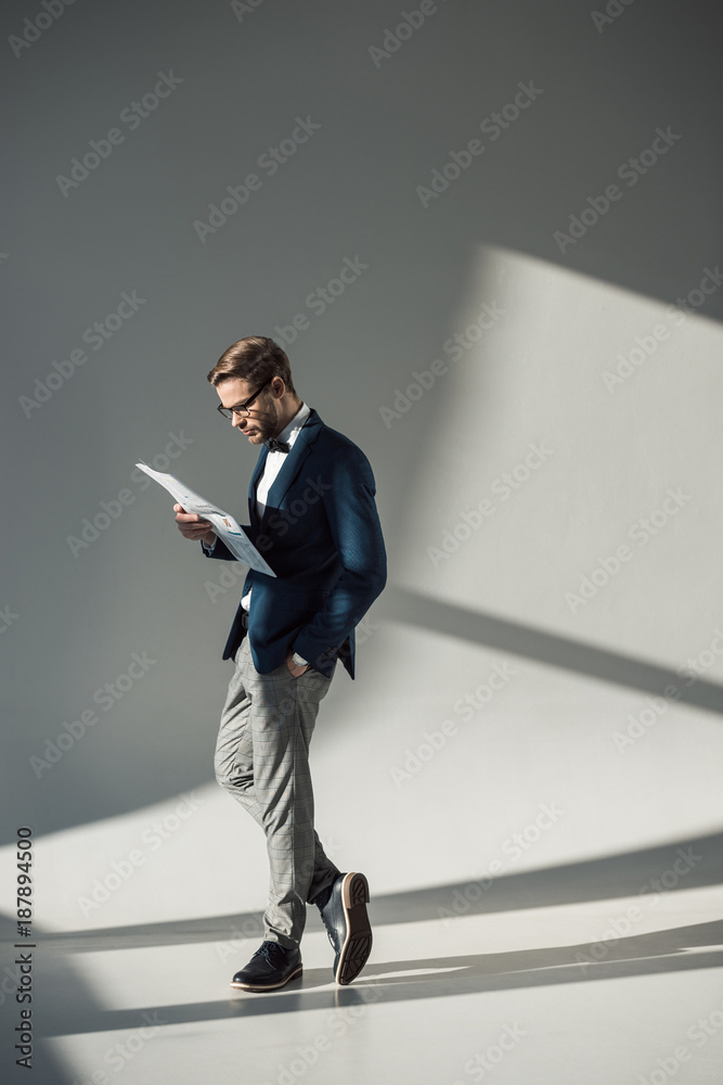full length view of handsome stylish businessman in spectacles reading newspaper on grey full length view of handsome stylish businessman in spectacles reading newspaper on grey