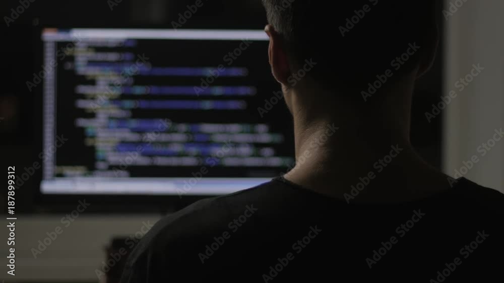 programmer from behind and programming code on computer monitor at night in a dark room