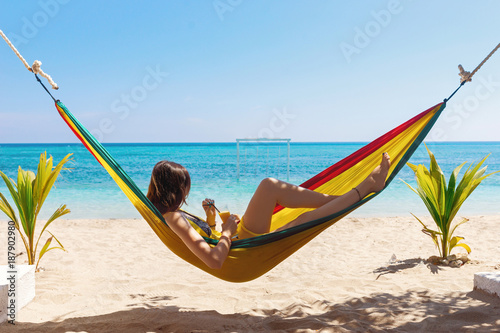 Girl with a glass of juice in her hand lies in a colorful hammock against the blue sea.Relaxing young woman in a hammock on the background of the sea Indonesia Bali Gili Air