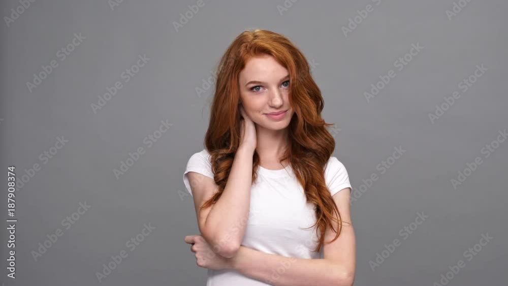 Embressed ginger woman in t-shirt with crossed arms touching her hair and looking at the camera over gray background Embressed ginger woman in t-shirt with crossed arms touching her hair and looking at the camera over gray background