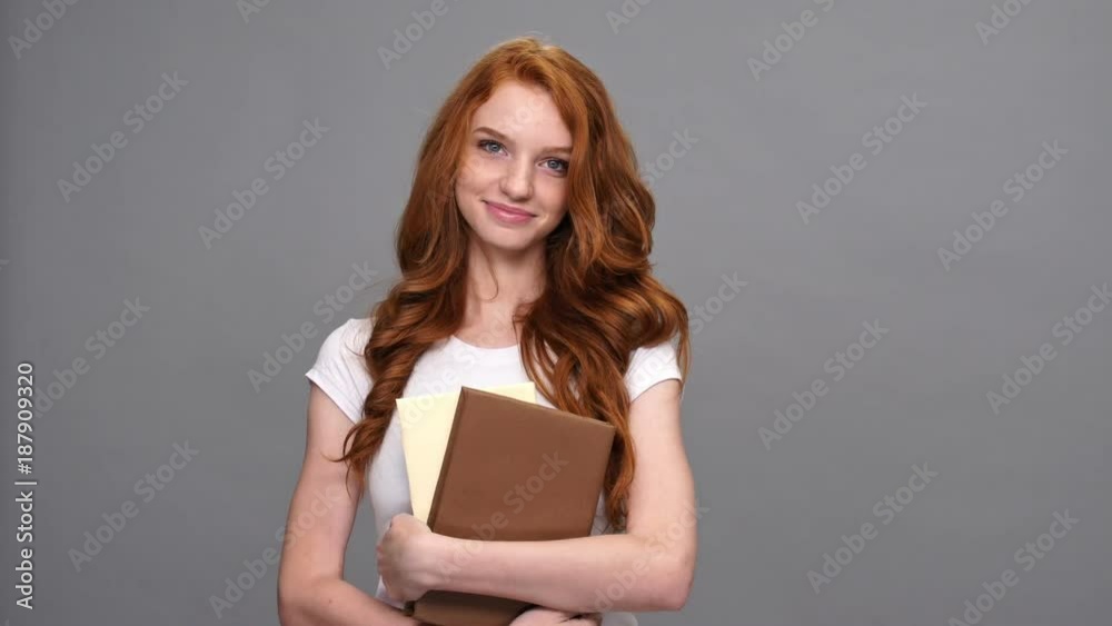 Embressed smiling ginger woman in t-shirt holding books and looking around over gray background Embressed smiling ginger woman in t-shirt holding books and looking around over gray background