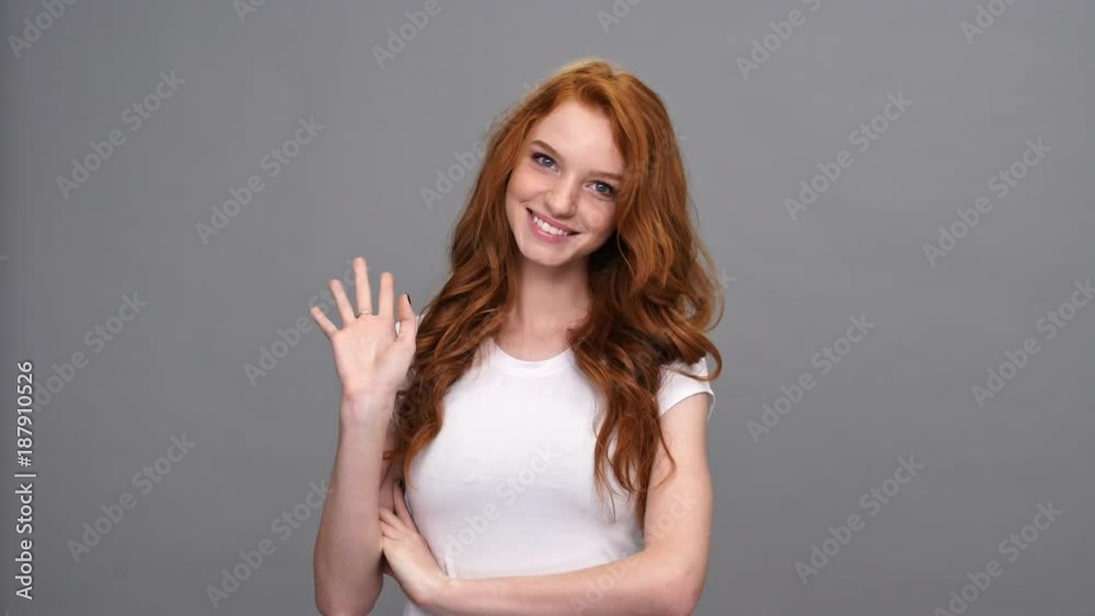 Smiling ginger woman in t-shirt holding crossed arms and waving at the camera over gray background Smiling ginger woman in t-shirt holding crossed arms and waving at the camera over gray background
