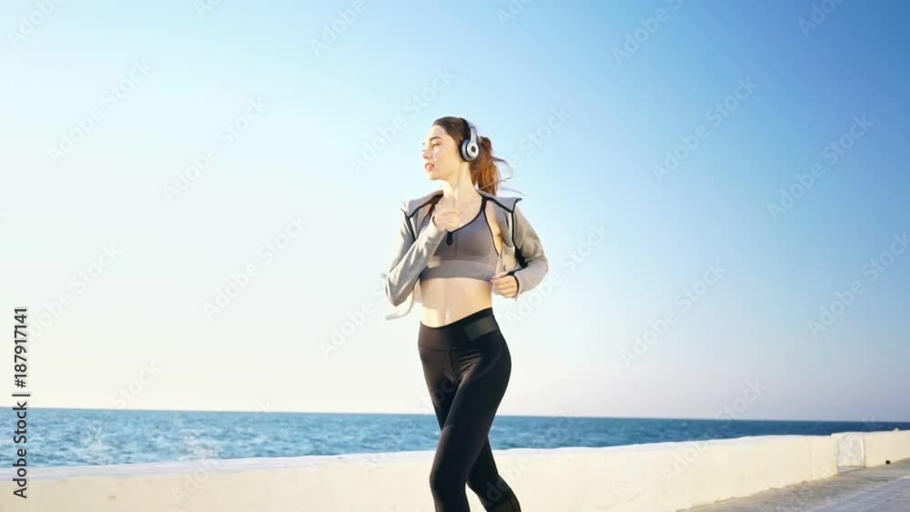 Full length view of Pretty brunette sports woman in headphones running at the seaside Full length view of Pretty brunette sports woman in headphones running at the seaside
