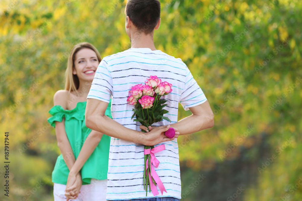 Young man hiding flowers and box with engagement ring before making proposal to his girlfriend in park Young man hiding flowers and box with engagement ring before making proposal to his girlfriend in park