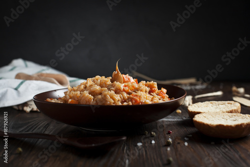 pilau in a brown ceramic dish, a kitchen towel, cherry tomatoes and a basket with spikelets of wheat, bread and a wooden spoon on a dark wooden table