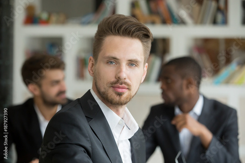 Young serious businessman looking at camera on corporate meeting with partners, professional confident company ceo boss leader manager co-owner in suit with diverse executive team, headshot portrait