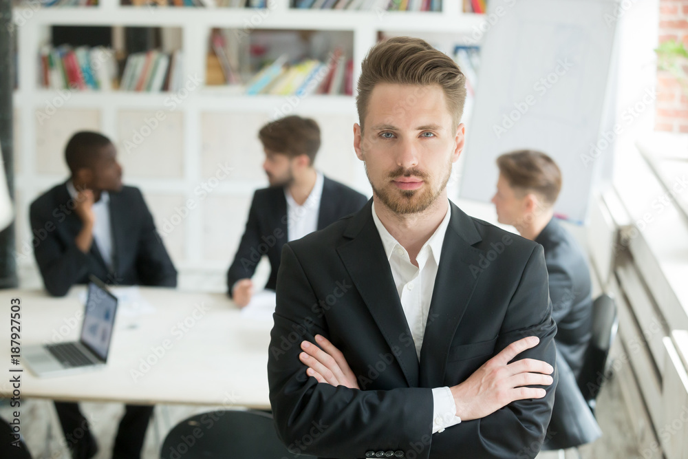 Serious young leader in suit looking at camera standing arms crossed on executive team meeting background, confident manager boss ceo co owner posing in front of partners group, headshot portrait