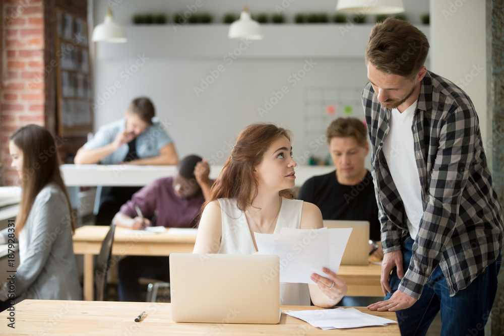 Female employee helping male colleague with documents in shared office, coworker explaining supporting new intern having questions about paperwork, assistance mentoring teamwork in co-working space