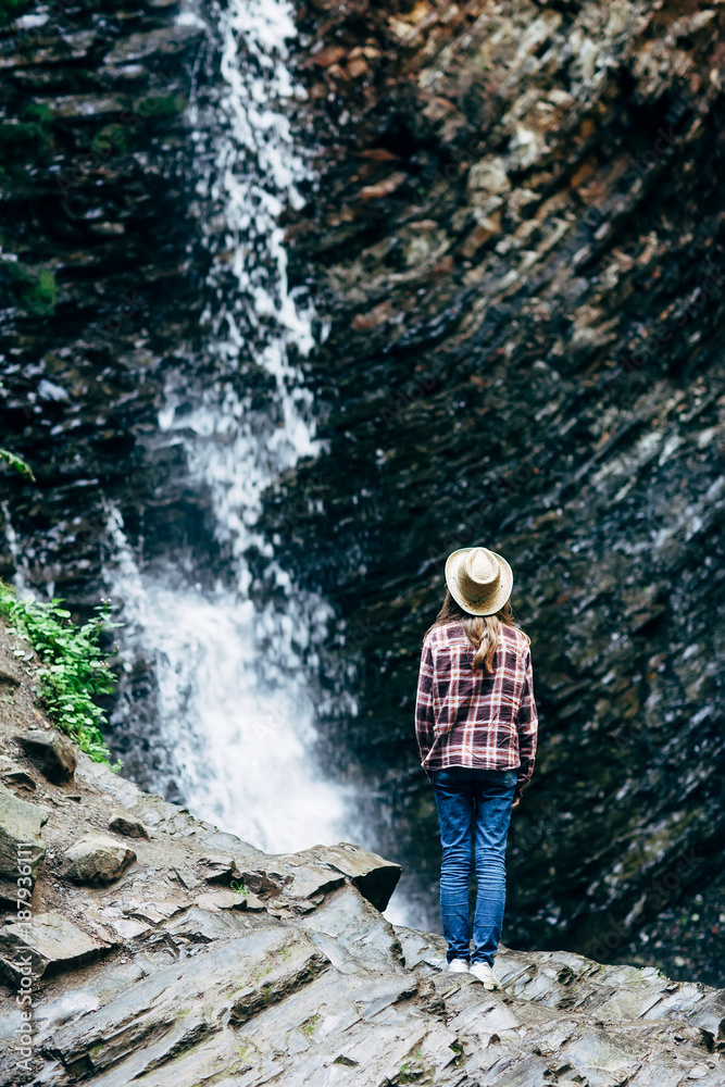 Girl in hat stands on rock and looks at waterfall