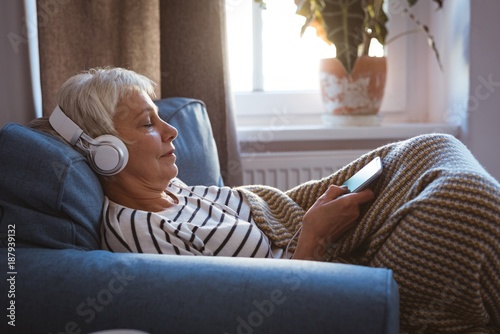 Senior woman listing to music on phone while siting on sofa in