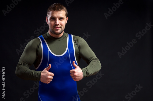 Young adult man wrestler in a blue tights shows thumbs up on a black isolated background