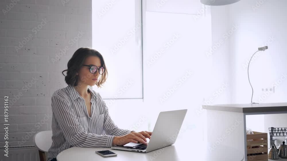Side view of concentrated brunette woman in shirt and eyeglasses sitting by the table and using laptop computer indoors

