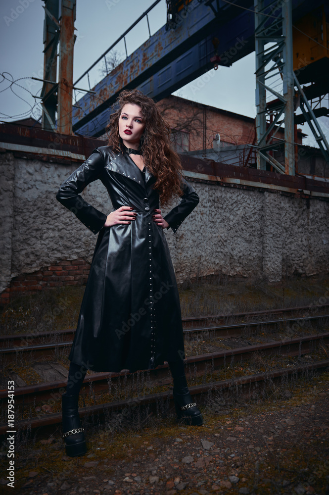 Fashion shot: portrait of beautiful goth girl (informal model) in leather coat standing at railroad (industrial area)