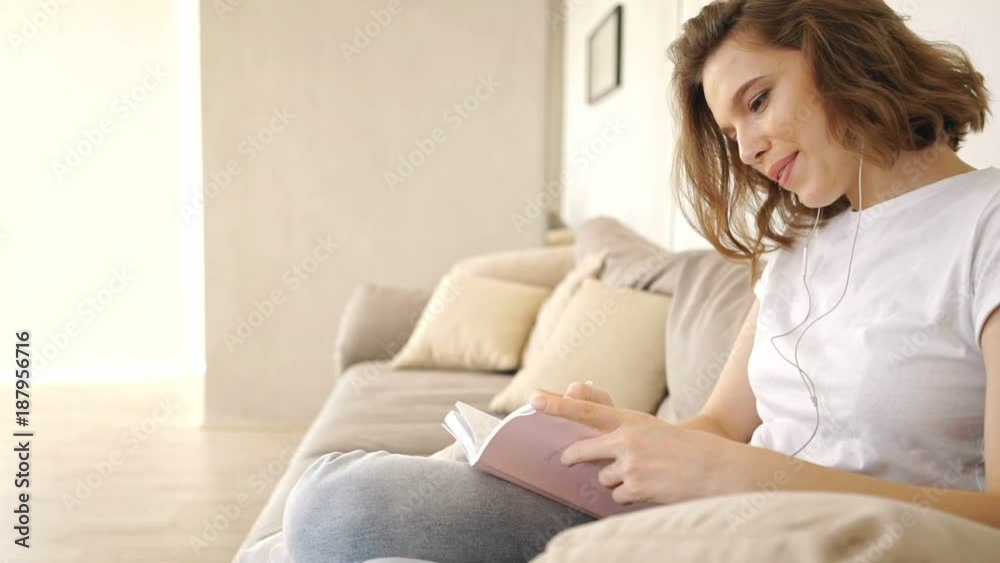Side view of Happy woman in earphones sitting on sofa and listening music while writing something in notebook at home Side view of Happy woman in earphones sitting on sofa and listening music while writing something in notebook at home
