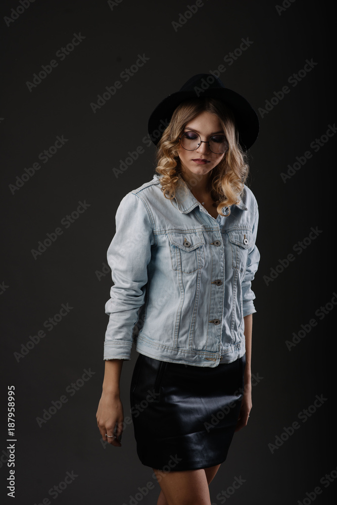 Fashion girl model posing on black background in the studio. Dressed in black hat, glasses and jeans jacket Fashion girl model posing on black background in the studio. Dressed in black hat, glasses and jeans jacket