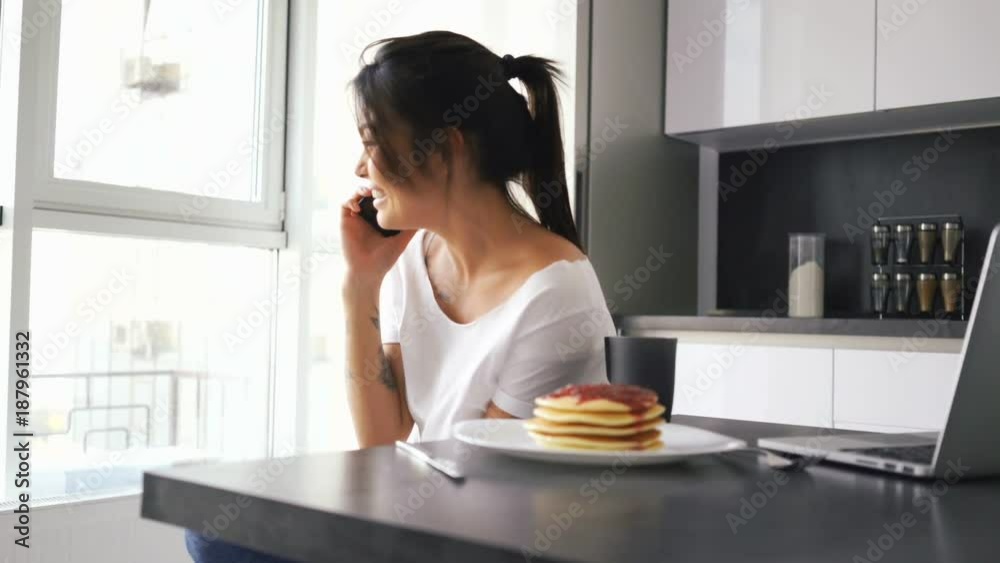 Happy asian woman sitting by the table on kitchen with laptop computer and pancakes while talking by smartphone and looking at window Happy asian woman sitting by the table on kitchen with laptop computer and pancakes while talking by smartphone and looking at window