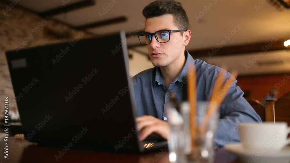 Handsome freelancer businessman in glasses diligently working on laptop in cafe. Man typing on keyboard and searches new job on internet at coffeeshop. Business concept