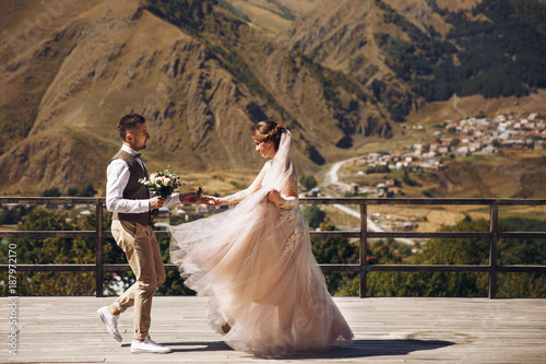 Groom in modern suit and bride in charming pink dress pose on the terasse with great mountain view in Georgia