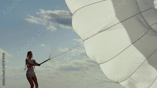 a young woman in modern and urban clothing drags a sail from a parachute to a sandy beach, in the background a clear sky with clouds, a lady with a stylish hairstyle makes an effort photo