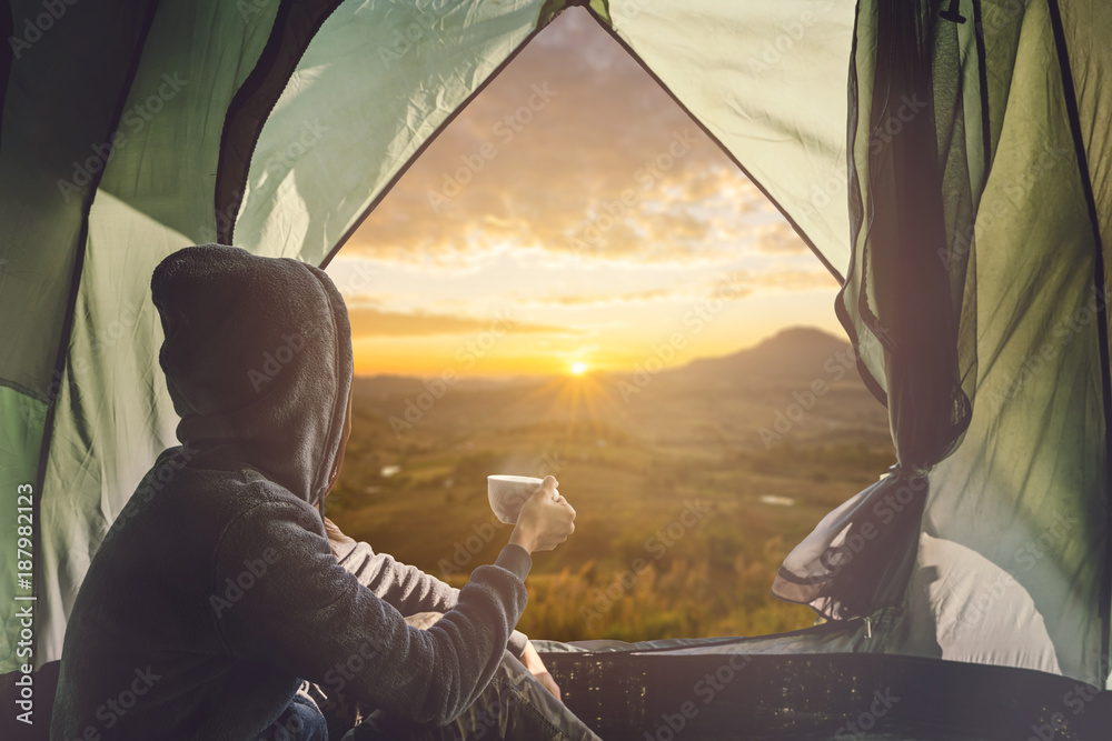 Young woman traveler sitting in the tent, relaxing and drinking coffee with beautiful sunrise - obrazy, fototapety, plakaty 