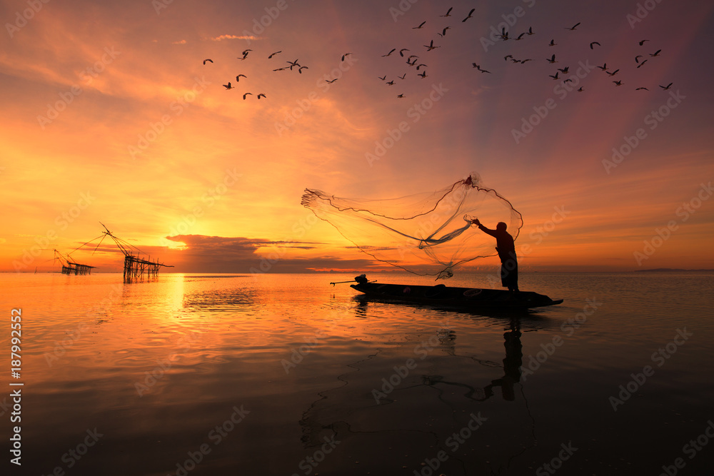 Asian fisherman on wooden boat casting a net for catching freshwater fish in nature river in the early morning before sunrise