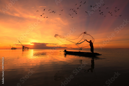 Fotografia Asian fisherman on wooden boat casting a net for catching freshwater fish in nat