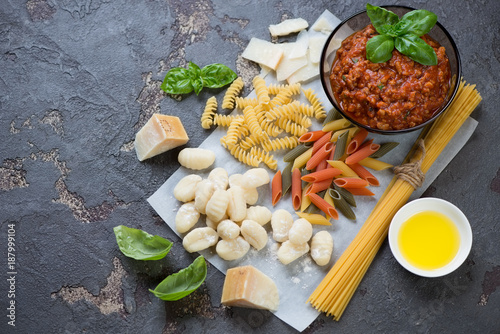 Different types of raw italian pasta with freshly made bolognese sauce. Elevated view on a brown stone background with space, horizontal shot