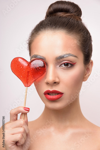 Beautiful young brunette woman. Fashion girl model with Valentine Heart shaped candy in her hand. Love Concept. Valentines Day gift. Red lipstick and arrow