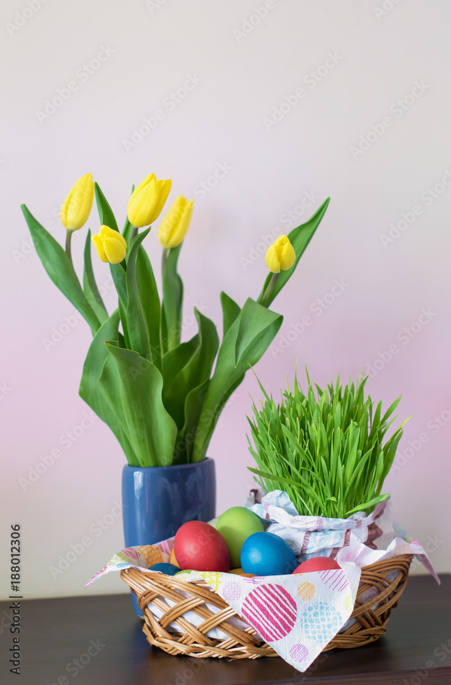A wicker basket with colorful Easter eggs, yellow tulips in vase, Easter decoration on light background. Free space for your text. Vertical image. A wicker basket with colorful Easter eggs, yellow tulips in vase, Easter decoration on light background. Free space for your text. Vertical image.