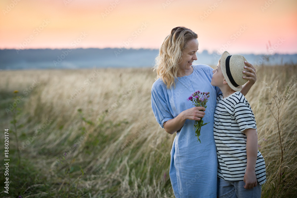 Portrait of happy family - mother and kid boy hugging while walking on the summer meadow at the sunset. Mom and child. Togetherness.