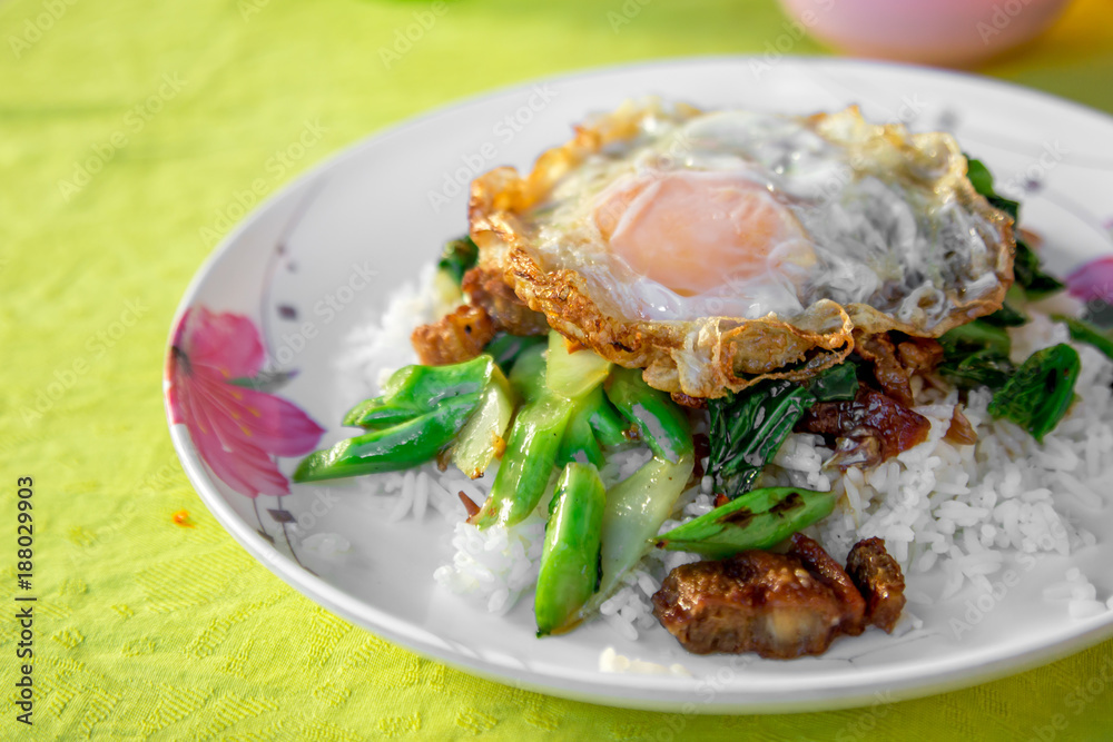 The fried crispy pork with fresh green kale and fried egg with oyster sauce and white rice in the white plate on the wooden table. The fried crispy pork with fresh green kale and fried egg with oyster sauce and white rice in the white plate on the wooden table.