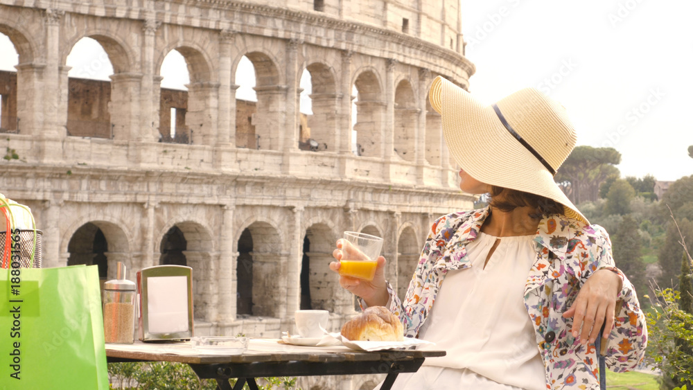 Happy young woman tourist drinking coffee and juice with cornetto at the table outside a bar restaurant in front of the Colosseum in Rome. Elegant beautiful dress with large hat and colorful shopping