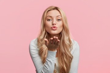 Beautiful woman with makeup and long blonde hair blows kiss, demonstrates her good feelings, says goodbye on distance, isolated over pink background. Young pretty female model makes air kiss.