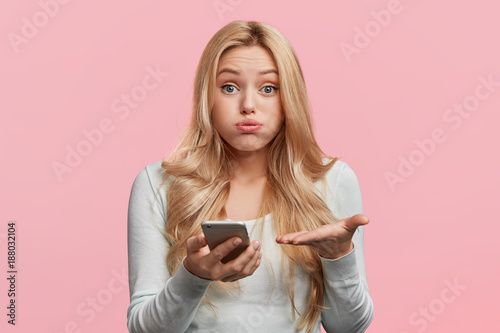 Indoor shot of indignant puzzled woman blows cheeks as looks into screen of modern cell phone, can`t understand content of recieved message, has frustrated look. Technology and people concept