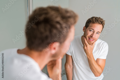 Male beauty young man touching beard and face looking in mirror - healthy skin. Skincare in home bathroom. After shave men lifestyle shaving concept.