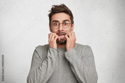 Tableau sur toile Stressful nervous bearded male bites nail with embarrassment, dressed in casual